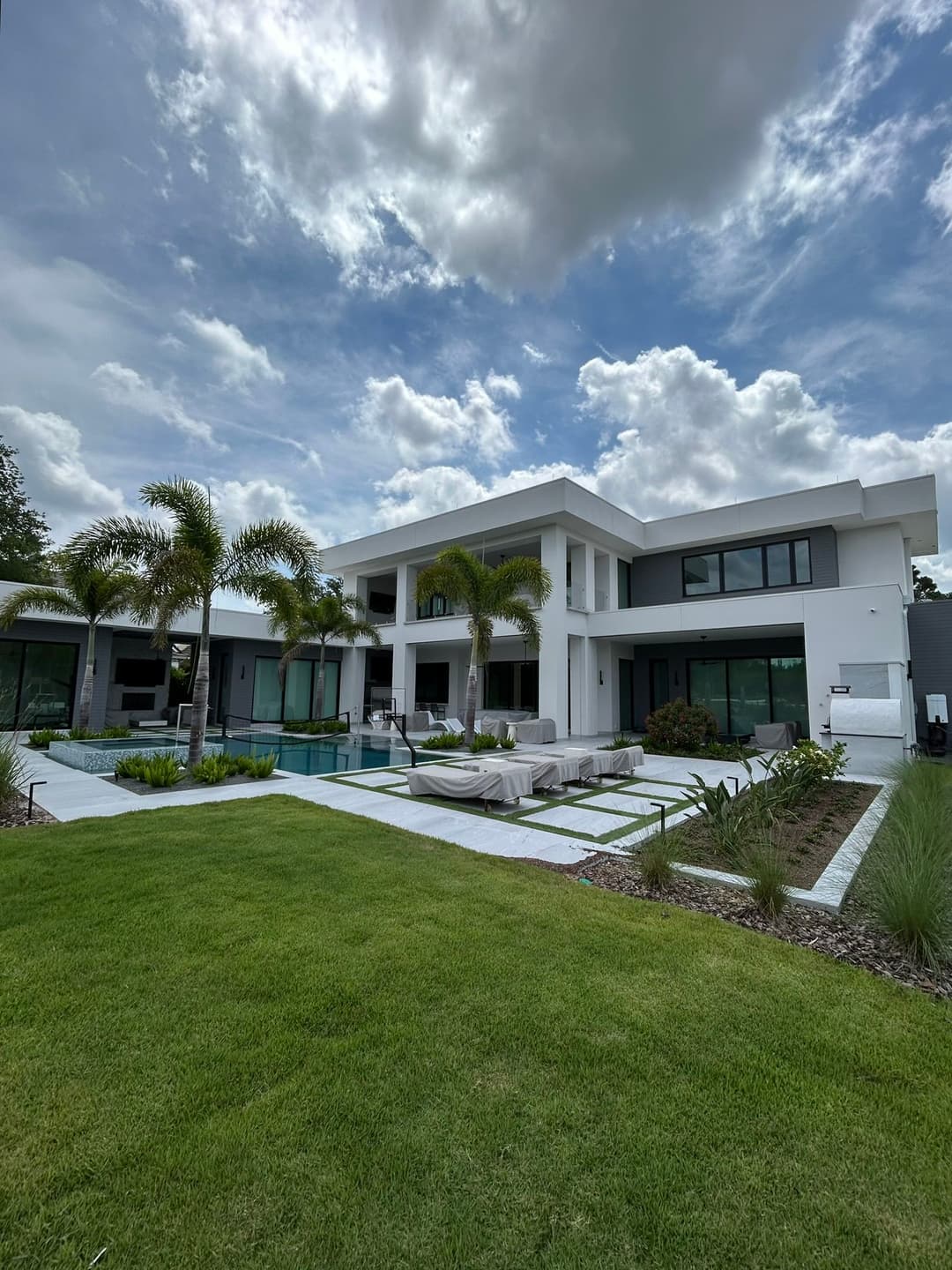 Modern luxury villa with palm trees, spacious patio, and a swimming pool under a cloudy sky.