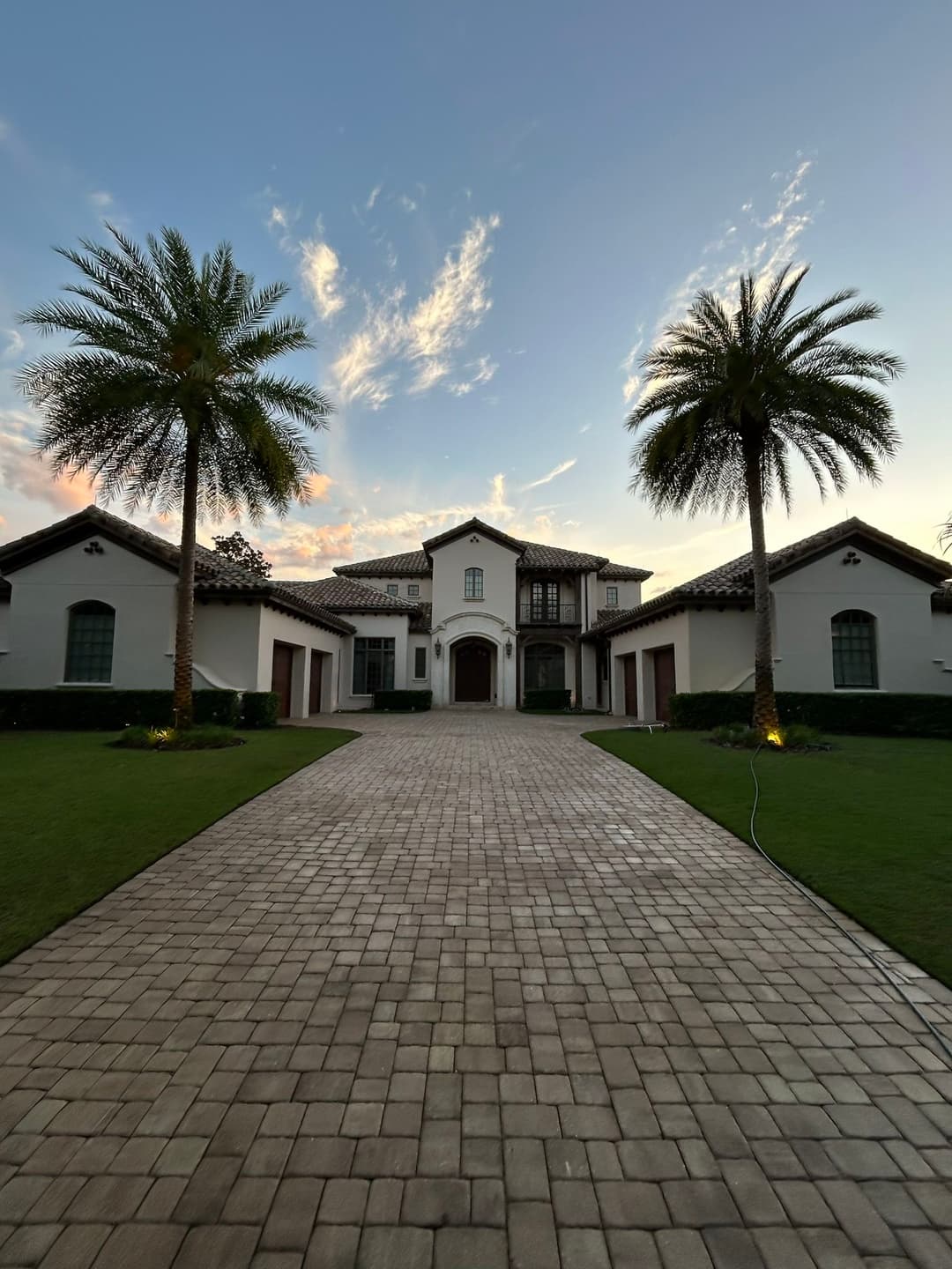 Elegant mansion entrance with palm trees and a beautiful sunset sky. Paved driveway leading up.