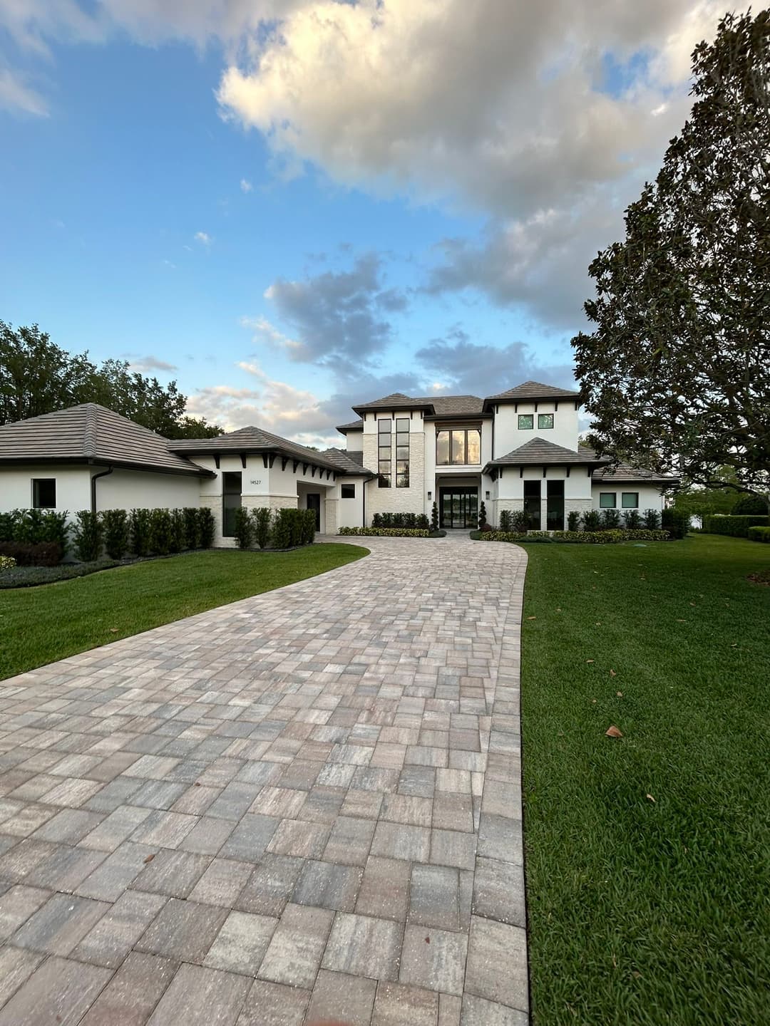 Modern luxury home with a brick driveway and lush green landscaping under a blue sky.
