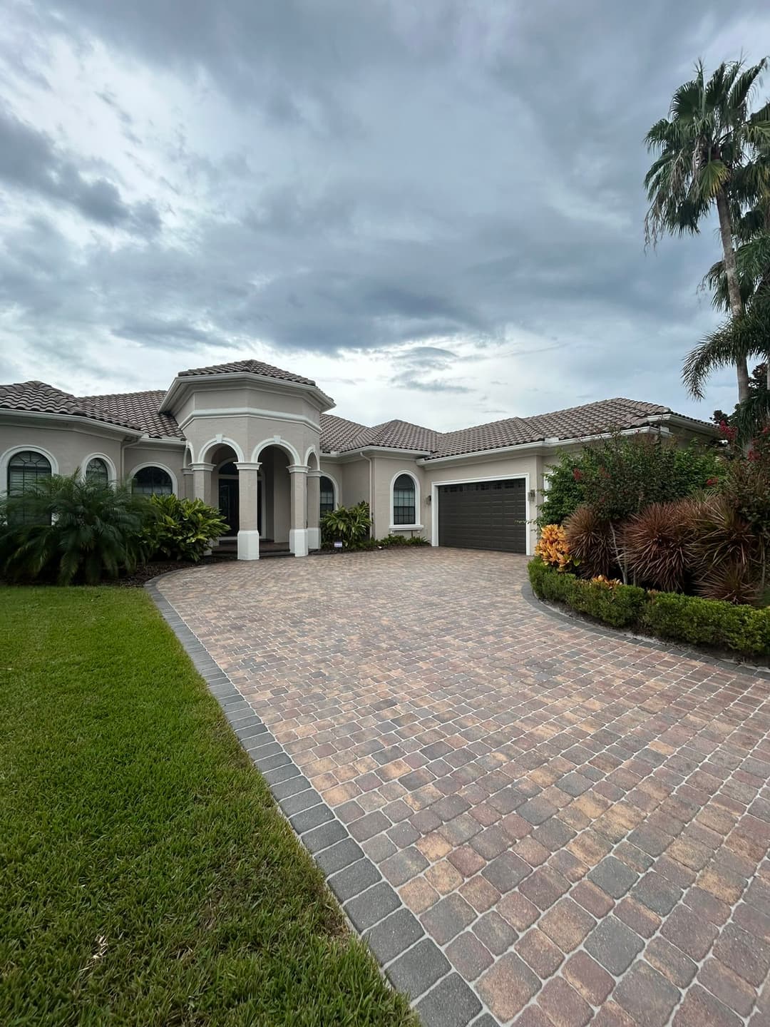 Elegant suburban home with a paver driveway, lush landscaping, and cloudy sky.